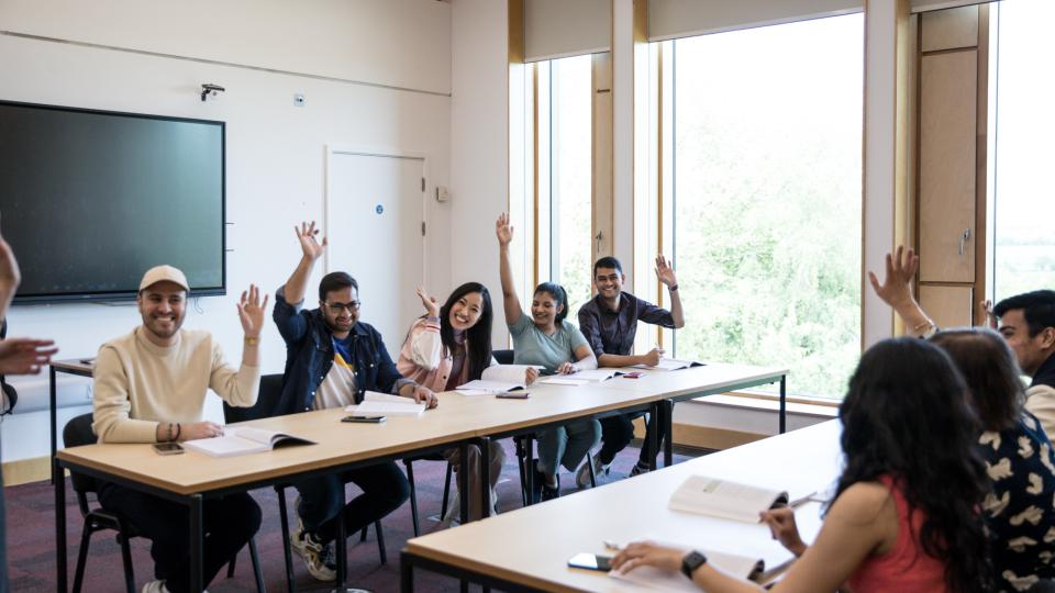 A group of students sitting at desks and working, raising their hands in a classroom setting.