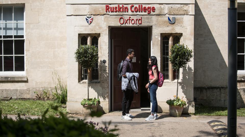 Two students standing, face-to-face, outside the main entrance to Ruskin College.