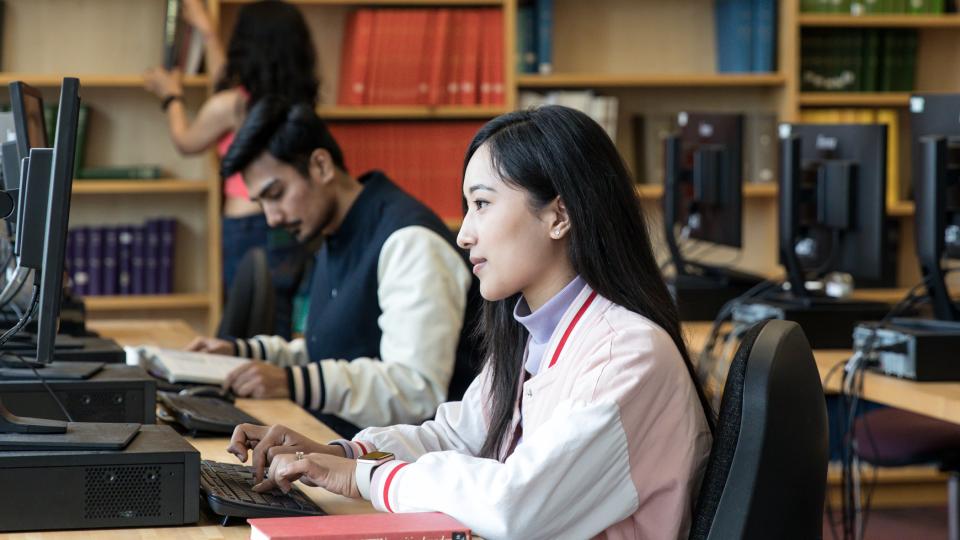 Two students working on computers in the library while another one is looking at books in the background.