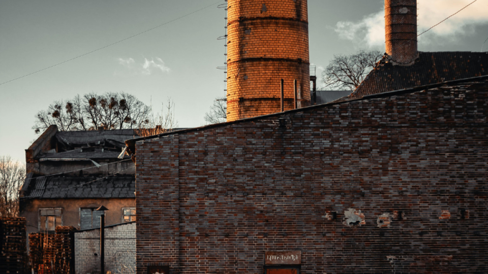 A brick structure which appears run-down is on a gravel track. Behind the brick wall is a tall chimney with a smaller one next to it.