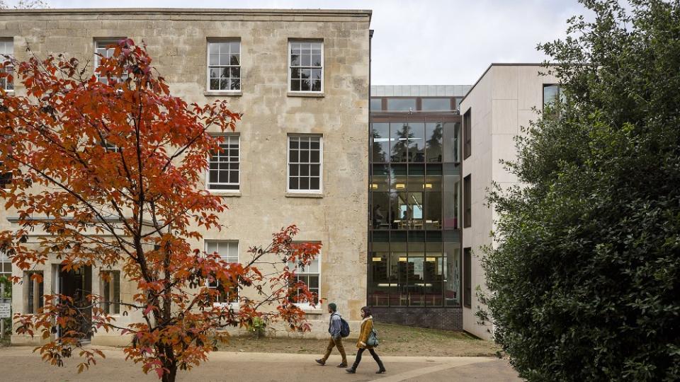 View of the outside of Ruskin college. Students are walking past the building and a tree has orange leaves.